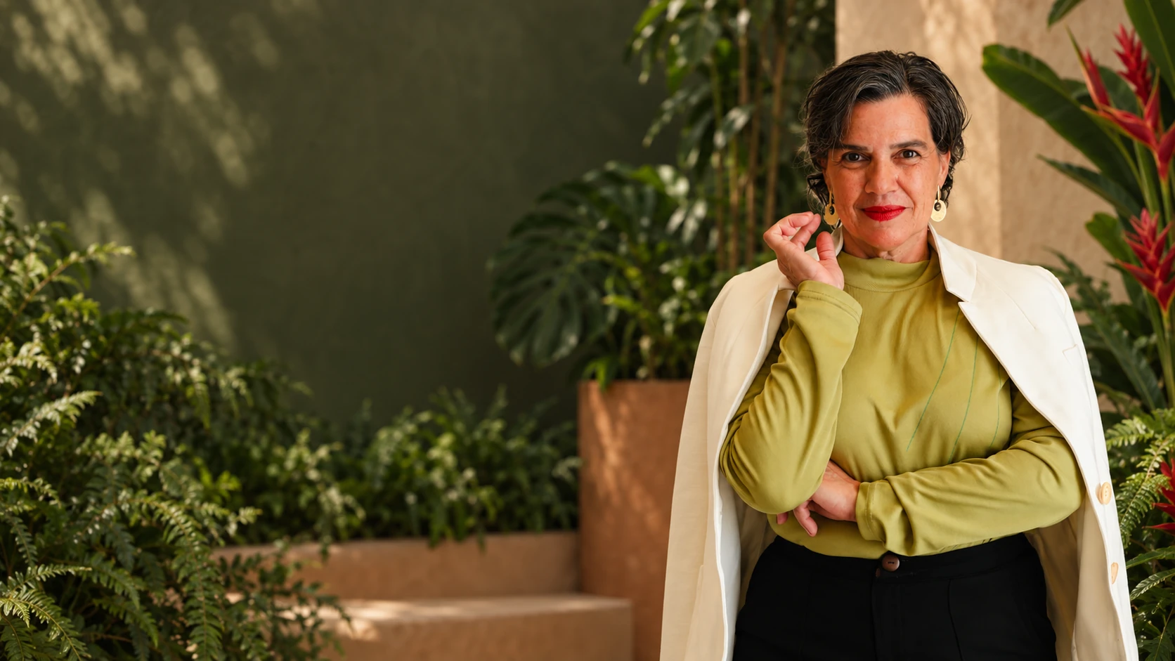 Valéria Pimentel, com blusa verde e blazer claro, em um cenário de plantas tropicais.