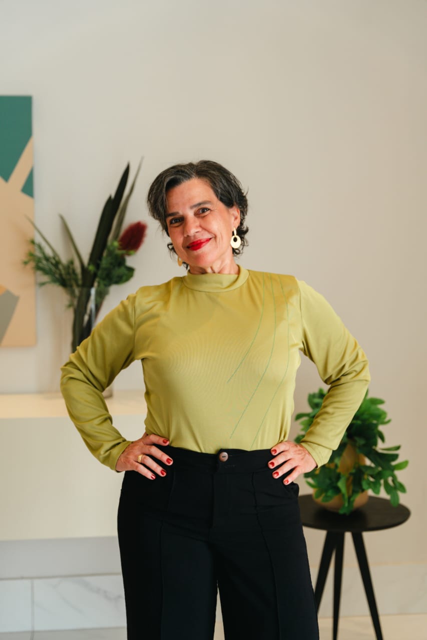 Valéria Pimentel sorrindo, com blusa verde e as mãos na cintura, junto a plantas em um ambiente de tons de areia.